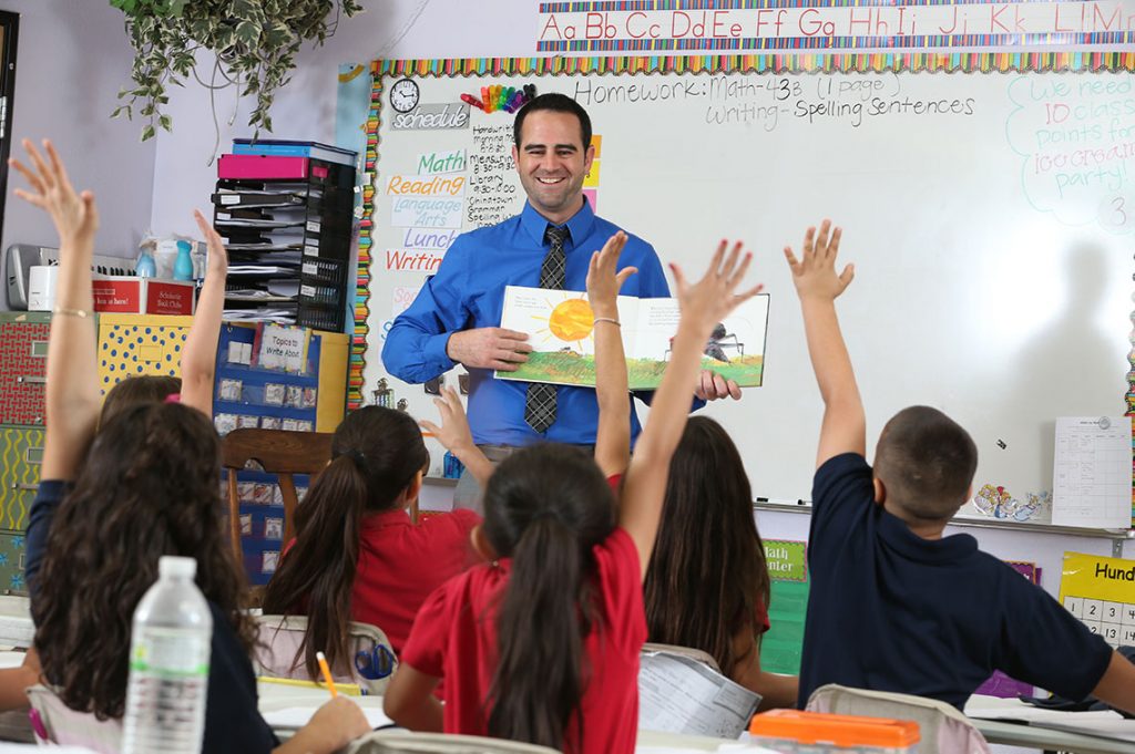 Male teacher reading in front of young students raising hands